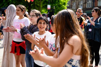 Children stand in a line outdoors with the sun shining. They are smiling and clapping hands.