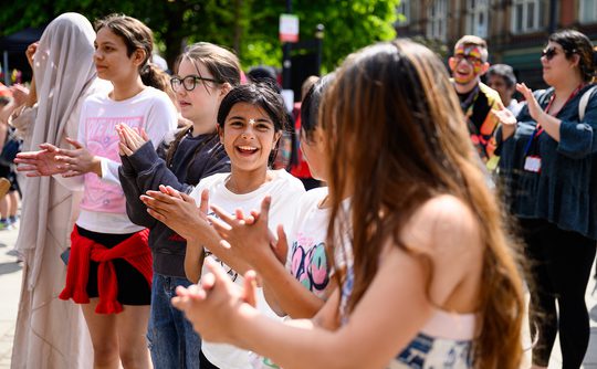 Children stand in a line outdoors with the sun shining. They are smiling and clapping hands.