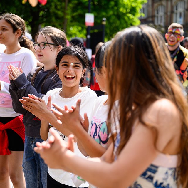 Children stand in a line outdoors with the sun shining. They are smiling and clapping hands.