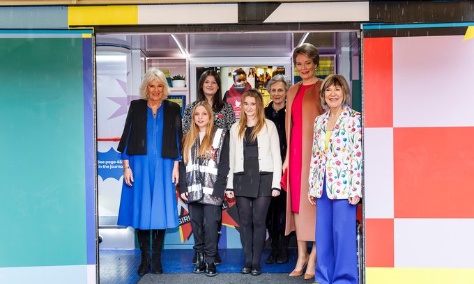 A group of people, including Her Majesty The Queen, Her Majesty Queen Mathilde of the Belgians, Her Royal Highness the Duchess of Gloucester & Jude Kelly, stand smiling inside a colourful, brightly-lit room. The atmosphere is cheerful and welcoming.