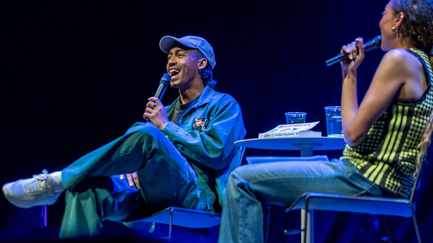 Two people sit on stage under blue lighting, holding microphones. Singer, Jordan Stephens is on the left, in a cap and blue outfit and appears to be laughing. The atmosphere is lively.