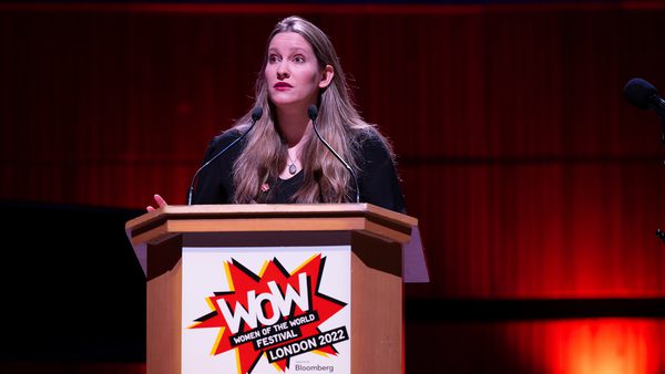 A woman speaks passionately at a podium labeled "Women of the World Festival London 2022." The stage is lit with warm red lighting,