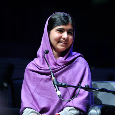 Young woman in a purple headscarf sits on stage, smiling warmly beside a microphone.