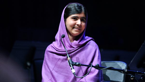 Young woman in a purple headscarf sits on stage, smiling warmly beside a microphone.