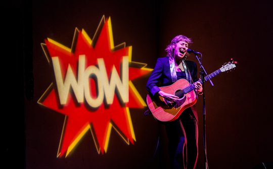 A musician passionately sings and plays guitar onstage, with a large "Wow" logo projected behind them