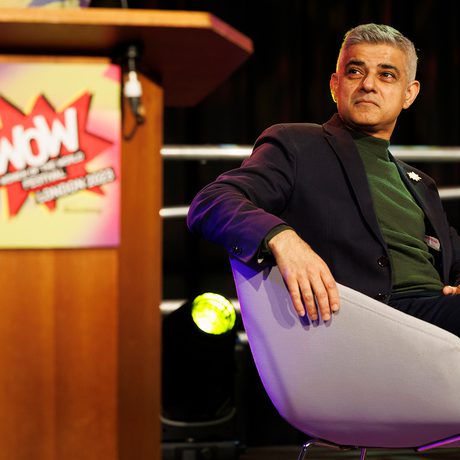 Sadiq Khan seated on a stage in a modern chair looks to the side, with a thoughtful expression. A podium with a "WoW" sign is in the foreground.
