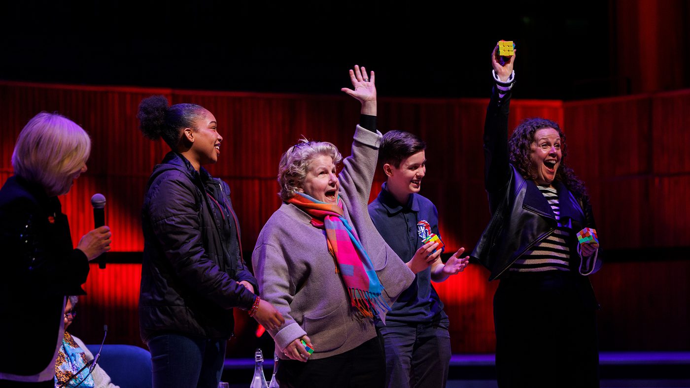 A group of four people stand on a stage behind Sandi Toksvig. Two people excitedly raise their hands, joyful expressions, in a warmly lit auditorium.