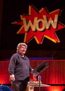 Sandi Toksvig smiling confidently on stage with a large red and yellow "WOW" sign projected behind them