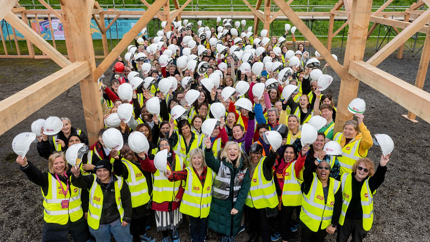 A large group of people in yellow vests and hard hats cheerily pose under a timber structure.