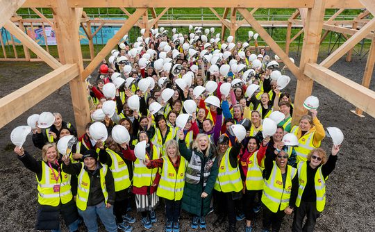 A large group of people in yellow vests and hard hats cheerily pose under a timber structure.