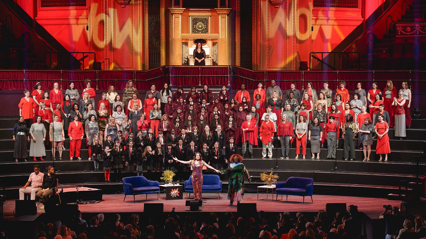 Concert in a grand hall with a large choir dressed in red, grey, and black. The lighting is vibrant and the audience is engaged