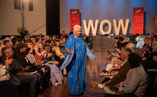 A woman in a vibrant blue dress walks confidently down an aisle between applauding audiences. The backdrop features large letters spelling "WOW."