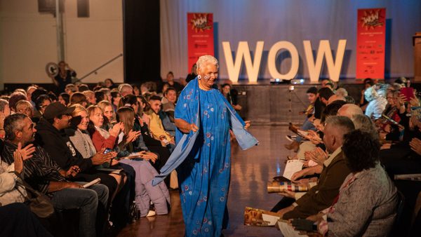 A woman in a vibrant blue dress walks confidently down an aisle between applauding audiences. The backdrop features large letters spelling "WOW."
