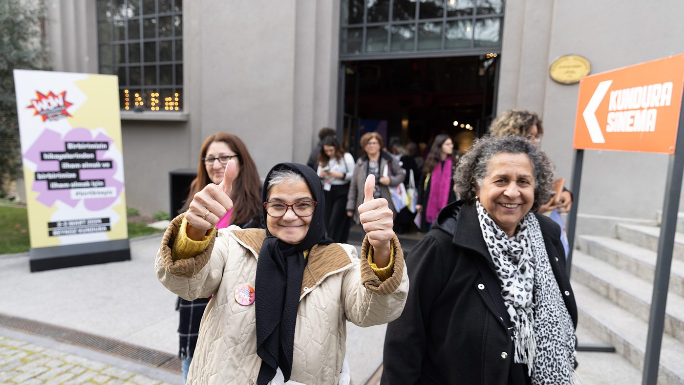 A group of smiling people exits a building, with a woman in front giving two thumbs up.