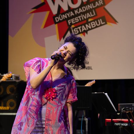 A woman performs passionately on stage, holding a microphone and wearing a vibrant dress. Musicians play instruments in the background. A festival banner hangs above.