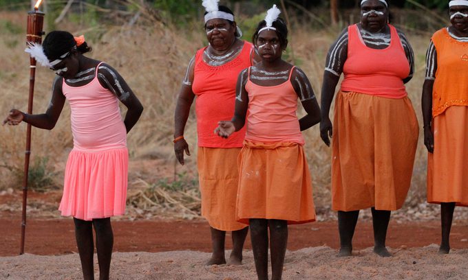 Five Indigenous women stand outdoors on a sandy area, wearing orange skirts and traditional body paint.