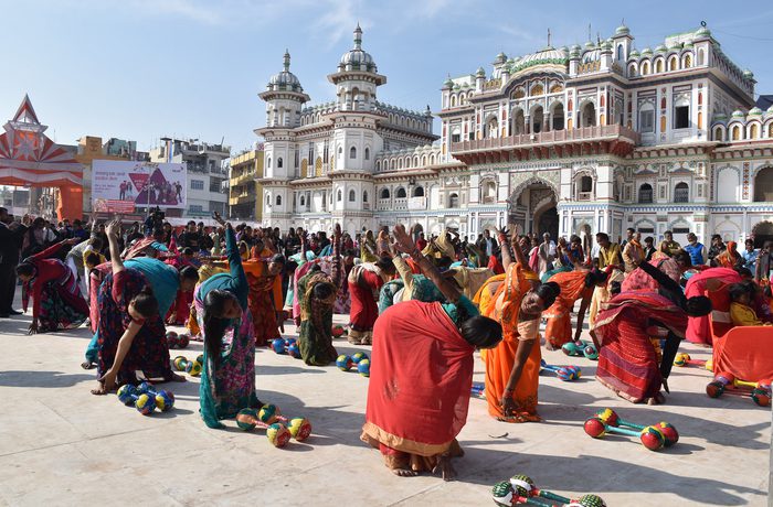 A large group of people in colourful attire perform yoga exercises outdoors. They are in front of a historical building. The scene is vibrant and lively.