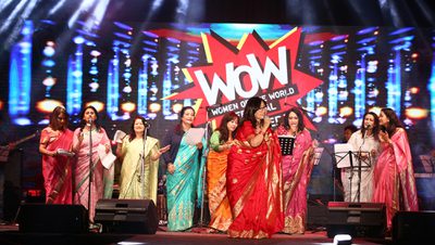 A group of women in colorful sarees on stage at the Women of the World event. Bright lights illuminate the scene, creating a joyful atmosphere.