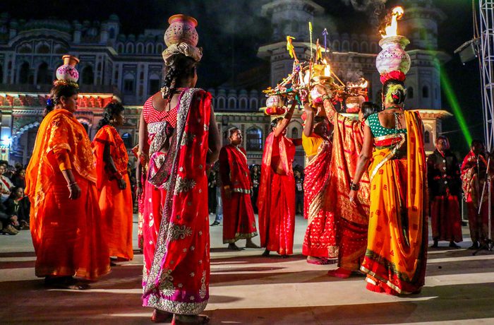 Women in vibrant red and orange sarees perform a dance at night, balancing decorated pots with flames on their heads.