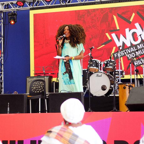 A woman speaks on stage at the WOW Festival Mulheres do Mundo in Rio. Bright decor, musical instruments, and an engaged audience set an inspiring scene.