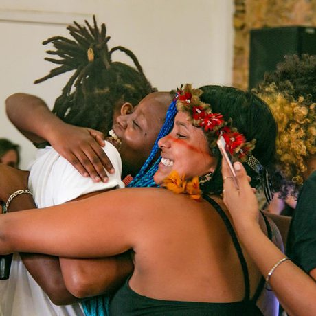 A joyful group hug in a lively setting. They are smiling warmly, with one wearing a floral crown, conveying happiness and camaraderie.