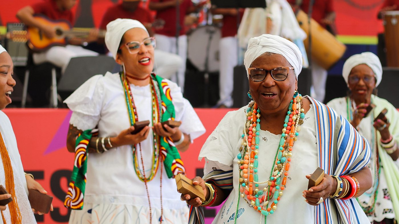 Elderly women in vibrant, traditional African clothing joyfully dance and play percussion instruments on stage with musicians in the background