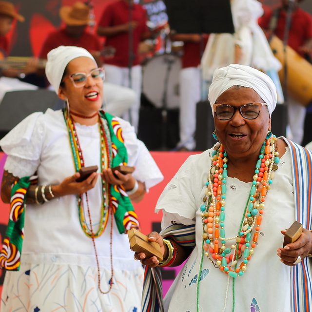 Elderly women in vibrant, traditional African clothing joyfully dance and play percussion instruments on stage with musicians in the background