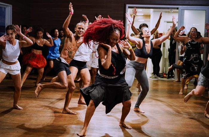 A group of people energetically dance in a studio, led by a woman with red hair in the centre. The room is filled with joyous expressions and lively movement.