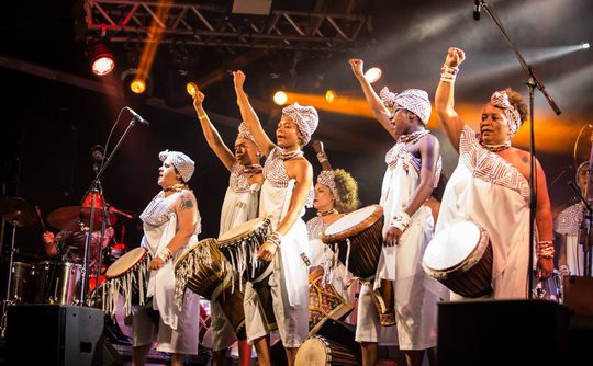 A group of women in traditional African style clothing attire play drums on stage under vibrant lights, raising fists in unity.