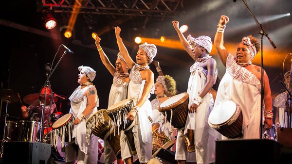A group of women in traditional African style clothing attire play drums on stage under vibrant lights, raising fists in unity.