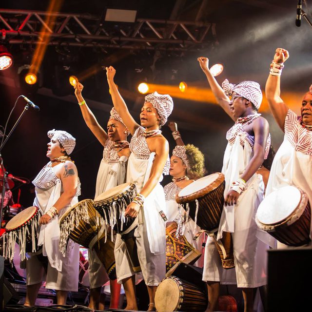A group of women in traditional African style clothing attire play drums on stage under vibrant lights, raising fists in unity.