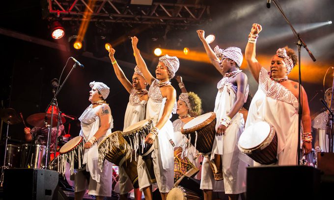 A group of women in traditional African style clothing attire play drums on stage under vibrant lights, raising fists in unity.