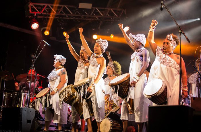 A group of women in traditional African style clothing attire play drums on stage under vibrant lights, raising fists in unity.