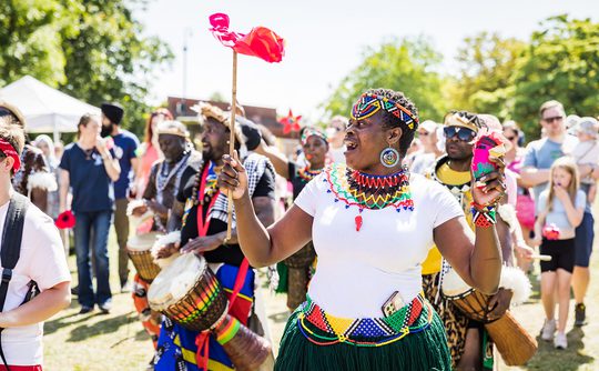 A joyful outdoor festival scene with a group wearing colourful attire, playing drums. The mood is vibrant and energetic, set against a sunny backdrop.