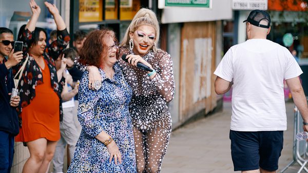 A drag performer in a sparkling outfit and a microphone joyfully embraces a smiling woman in a blue dress on a lively street, with cheering onlookers.