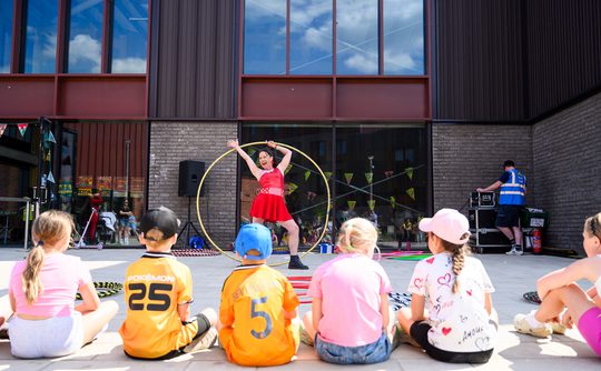 A performer in a red dress entertains a row of seated children with a large hoop outdoors. The backdrop is a modern building