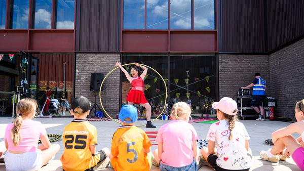 A performer in a red dress entertains a row of seated children with a large hoop outdoors. The backdrop is a modern building