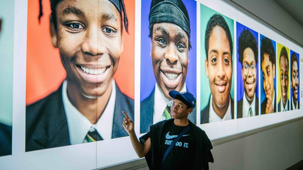 A young man stands by a wall of photos taken by Kay Rufai as part of his Smiling Boys Project. They are all photos of Black school boys smiling.